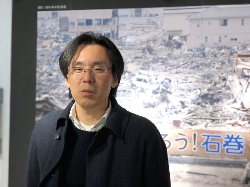 みらいサポート石巻専務理事、中川政治さん。初代の「がんばろう！石巻」看板の写真の前で。